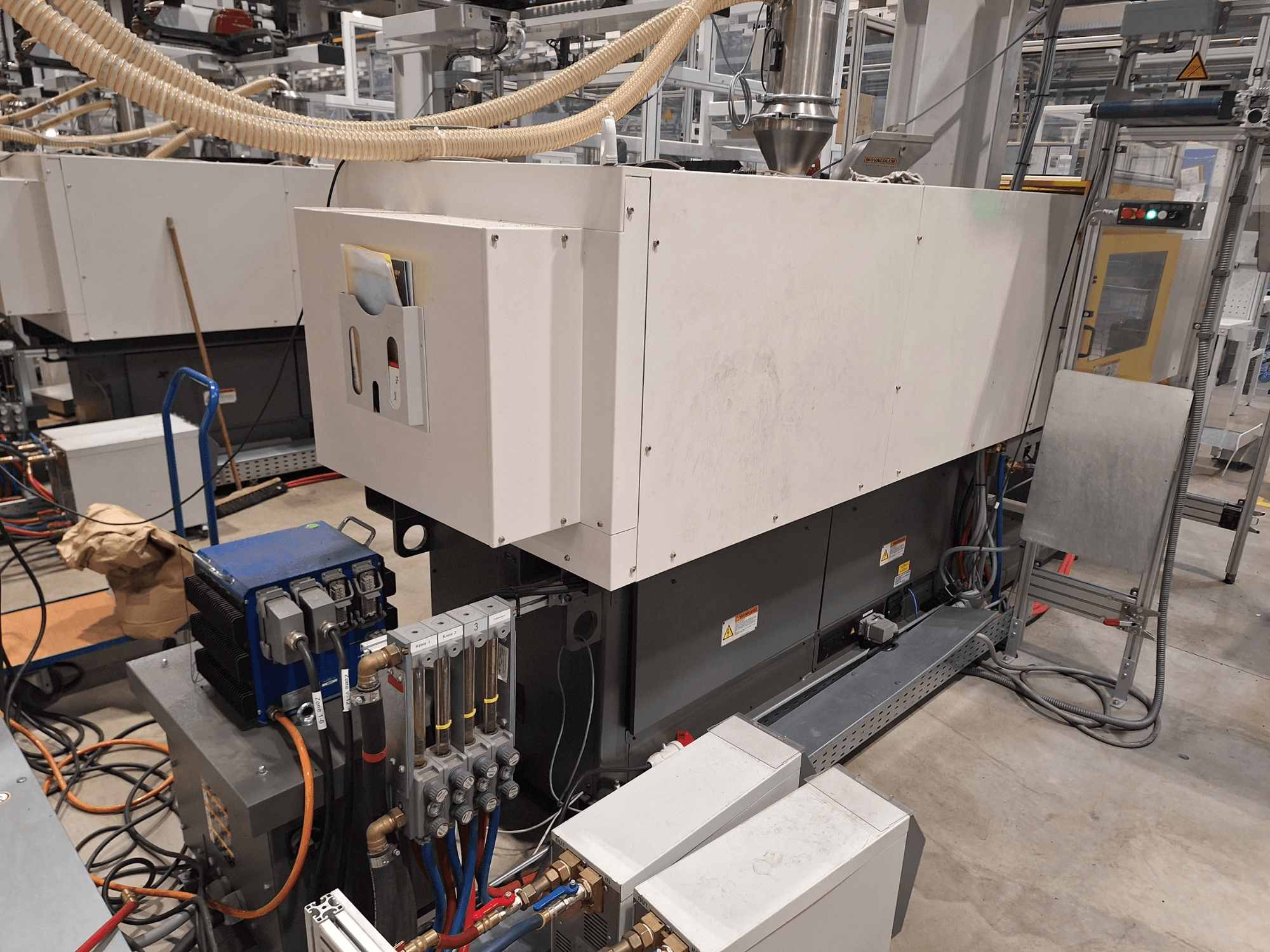 Industriell utrustning i en fabriksmiljö; inkluderar en stor maskin med kontrollpanel, slangar och elektriska anslutningar, vy från sidan.