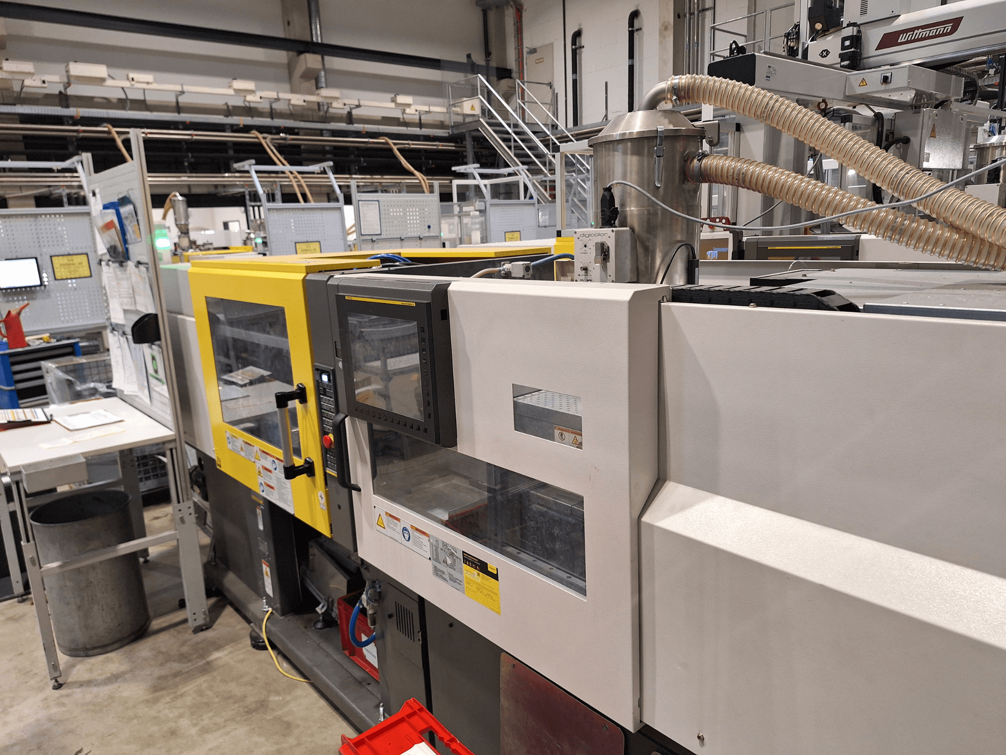 Frontvy av en flerfärgad formsprutningsmaskin med gula och grå sektioner, med kontrollpanel och avgassystem.