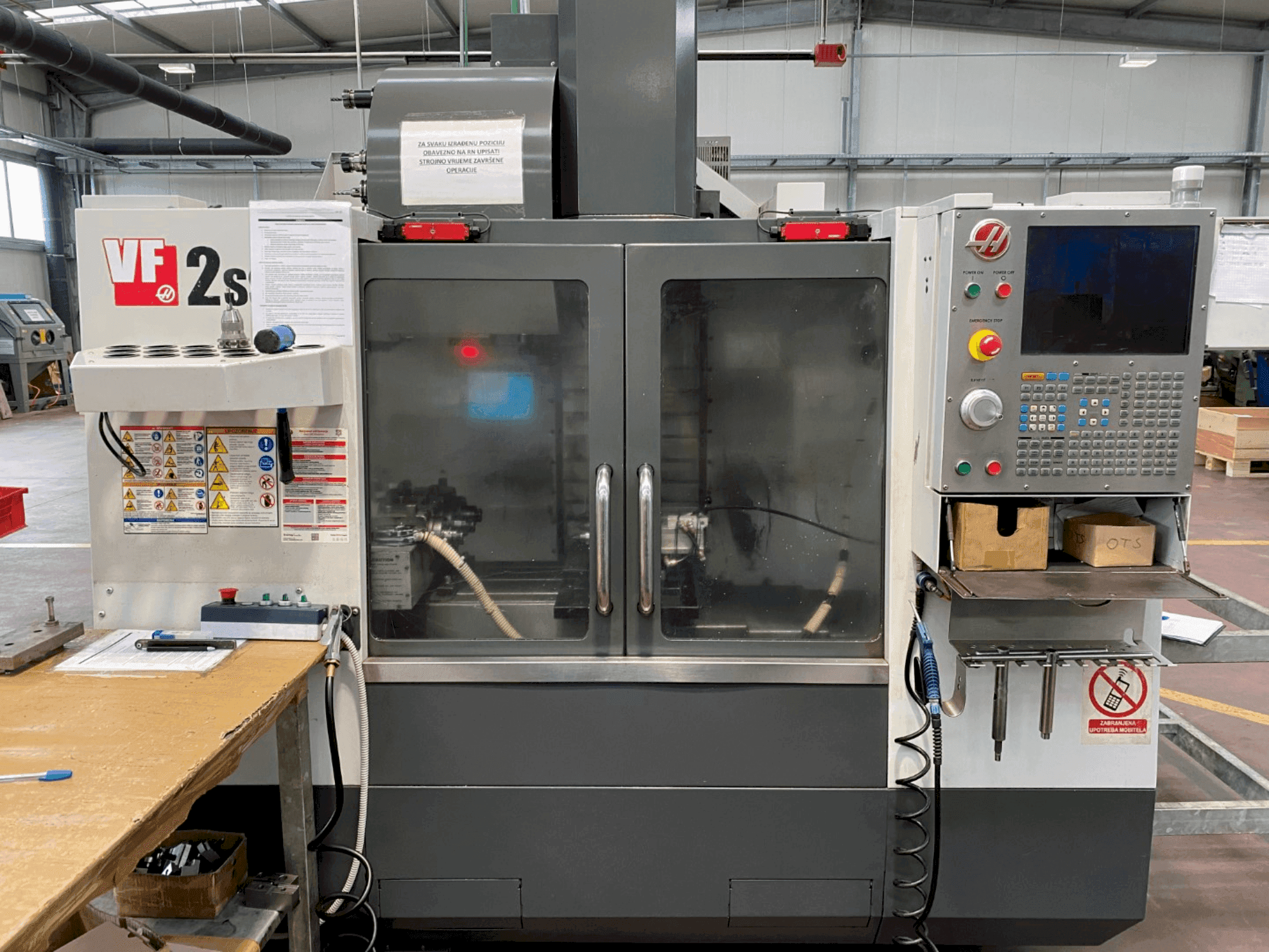 Haas VF-2SS bearbetningscenter framifrån, med kontrollpanel, kylvätskeslang och säkerhetsetiketter på sidan.
